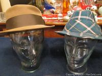 Two vintage hats on glass head forms, one tan fedora with brown ribbon, one plaid hat with a check pattern.