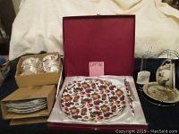 Photo showing full tea cup set with box, six cups and saucers stacked in boxes to the left, cake plate in satin-lined box center, metal stand with some cups and saucers displayed to the right.
