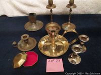 Full view of 8 brass candle holders of different styles and sizes, some with handles and some without. A hexagonal brass box with red interior is also present.