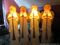 Four illuminated angel blowmold figures holding trumpets, glowing softly in a dimly lit room.