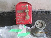 Vintage red fire alarm pull box and brass fire hose nozzle placed on ground; fire alarm shows lever and label.