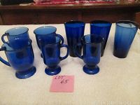 Seven blue glasses arranged on a soft white surface. Three have handles and are shorter, four are taller without handles.
