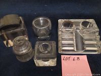 Photo shows a group of six ink wells, mostly clear glass in various shapes including square, round, cube-shaped, and multi-pen rest style. Sizes are approximately 2-3 inches.