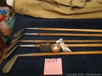 Wide shot showing multiple vintage wood shaft golf clubs laid horizontally on a dark surface, with a canvas golf bag partially visible above them and a pink lot tag labeled 'LOT 69'