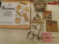 Photo showing the Cardinal Teasers box stating 7 different brain busters crafted of solid wood, alongside various wooden and metal brain teaser puzzles arranged on a cloth surface.