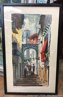 Framed watercolour painting showing full street scene with person and donkey below an archway and buildings with red awning and shutters.