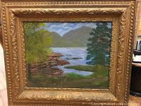 Full view of the oil painting displayed in its frame showing the landscape with a lake, trees, rocks, and hills.