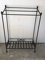 Front view of a metal quilt/blanket rack showing three horizontal rods on top and a lower slatted shelf, with round finials on the vertical posts.