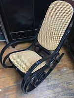 Front angle showing the black bentwood frame, cane seat and backrest, and wooden floor beneath.