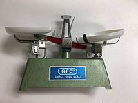 Front view of the GFC Small Gold Scale showing the green metal base, two white pans, and balancing beam with a red accent. The blue and white GFC label is visible on the front.
