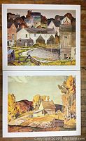 Two unframed A.J. Casson prints depicting village and rural autumn landscape scenes, both measuring 24 x 20 inches.