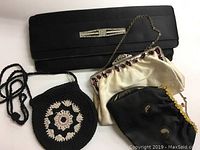 Four vintage purses arranged on a white surface, showing a black satin clutch, cream satin purse, black fabric purse, and black crocheted purse with floral design.