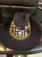 Front angle showing part of the collection of vintage pins on the black cowboy hat and the gold rope trim.