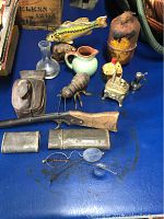Photo showing vintage eyeglasses in metal cases, small antique rifle, various ceramic pitchers, metal insect figurine, and collectible figurines on blue surface.