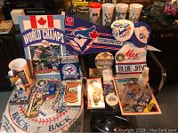 Full view of the lot showing various Toronto Blue Jays items including cups, mugs, pins, pennants, commemorative plates, a bobblehead, and collectible cards focused on 1992 World Series.