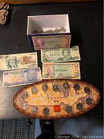 1999 Millennium Canada coin set displayed on an oval wooden map plaque with multiple coins, several world paper bills next to the plaque, and a white box containing loose coins.