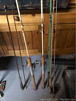 Five vintage fishing rods standing against a cabinet, showing cork handles and two rods with reels mounted