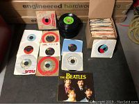 Photo showing a collection of approximately 45 RPM records arranged with some in paper sleeves, one Beatles record with illustrated sleeve visible, and a box with more records inside.