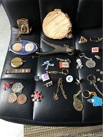 Image shows an assorted collection of vintage collectibles arranged on a black leather surface. Items include various metal pins, medallions, two coins, a wooden slice, a metal clamp tool, a round brass pendulum, and other small vintage items.