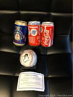 View of signed baseball with attached photo sticker, certificate of authenticity and three collectible tins arranged behind on a black surface