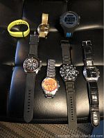 Seven assorted watches displayed on black surface including chronograph, sport digital, vintage and fitness tracker styles.