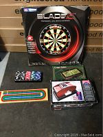 Photo of the entire lot including boxed Winmau Blade 5 dart board, card shuffler box, mini craps table, poker chips, and cribbage board.