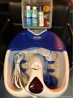 Dr. Scholl's foot bath massager with power cord and three rolling attachments placed on top, alongside a clear box containing multiple small bottles and tins likely of balms and natural mouthwash.