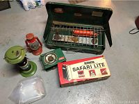 Photo shows the green Coleman mini burner stove with metal case, two vintage lanterns (one red and one green), the Burgess Safari Lite fluorescent light in its box, and a clear collapsible water bag.