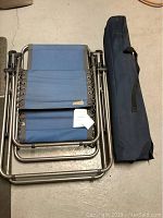 Folded blue camping cot with attached paper tag beside navy blue carrying bag with handles, placed on concrete floor.