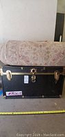 Photo showing black steamer trunk with brass hardware and a rolled-up rug on top