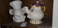Photo showing a white ceramic cookie jar shaped like Mickey Mouse wearing a chef hat next to a colorful teapot-shaped cookie jar resembling Mrs. Potts from Beauty and the Beast.