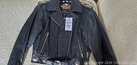 Front view of small black leather women's Harley Davidson biker jacket with zipper pockets and silver concho studs on belt and epaulets.