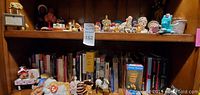 Overview of shelf with small collectible figurines and plush toys including Ty Beanie Babies, various animal and human figurines, and books in background