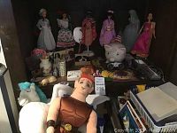 Barbie dolls in various traditional costumes alongside a large plush winged character doll and a white unicorn toy on a shelf.
