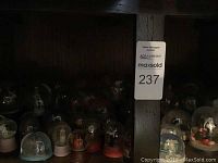 Photo showing multiple snow globes on wooden shelves, with some bases in red, white, blue, and black and various figurines inside glass domes.