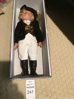 Doll inside white box dressed in black jacket, brown patterned vest, white pants and black boots.