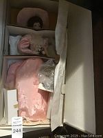 Porcelain doll dressed in pink gown with bonnet, inside box with tissue wrapping and accessories in plastic bag.