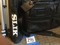 Photo showing black leather Tumi bag side view and one tripod in silver with black soft carrying case labeled SLIX.