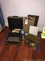 Full view of vintage Remington Portable typewriter in case with clipboards, office supplies, and storage box arranged on hardwood floor.