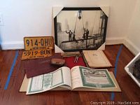 Photo shows three vintage New York license plates, framed photo, books, drafting tools and broken spectacles laid out on floor.