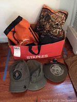 Two military-style hats, a hiking canteen and three tote bags displayed on floor with box.