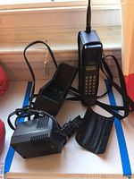 Photo showing the Radio Shack cellular phone standing upright with extended antenna, accompanied by its charging dock and Archer AC adapter cables.