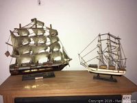 Photo showing two wooden model ships displayed on a wooden surface against a plain wall.