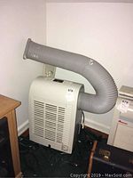 White Friedrich portable air conditioner with flexible exhaust hose attached, positioned on floor near corner.