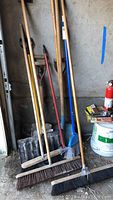 Full view of 11 large garden tools including shovels and various brooms piled together showing handles and working ends, situated in a corner with some paint buckets nearby.
