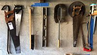 9 vintage hand tools hanging on wall, including saws, crowbars, level, and brushes