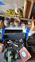 Photo showing open vintage metal toolbox with numerous hand tools and hardware inside, rolls of tape and yellow rope nearby
