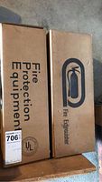 Two cardboard boxes side by side with labels indicating they each contain a fire extinguisher. Boxes marked 'Fire Protection Equipment' and 'Fire Extinguisher'.