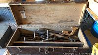Open wooden toolbox showing assorted vintage tools including a handsaw, brace drill, and wood plane.