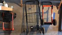 Photo showing two orange metal dollies and one large metal wire shopping cart against a wall.